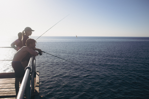 Serene silhouette of teenage children / siblings fishing together on jetty - Australian Stock Image