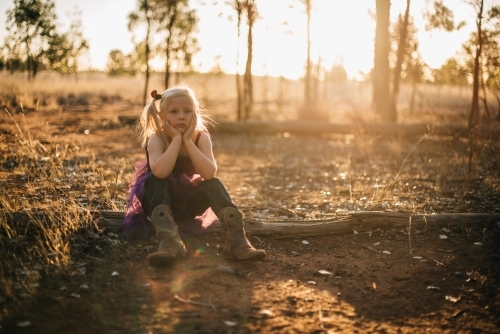 Serious young girl sitting outside with face leaning on hands as the sun sets - Australian Stock Image