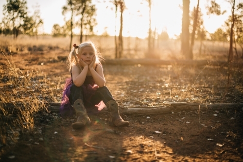 Serious young girl sitting outside with face leaning on hands as the sun sets