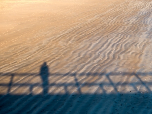 Shadow of a person, leaning on a rail, on a rippled beach sand pattern - Australian Stock Image