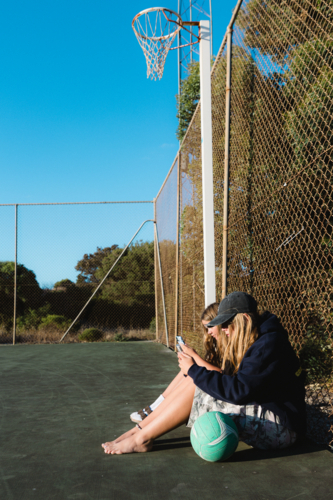Side image of teenage girls with ball on mobile phones at netball court - Australian Stock Image