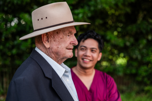Side portrait of an elderly man wearing a hat and suit outdoors assisted by male healthcare worker - Australian Stock Image