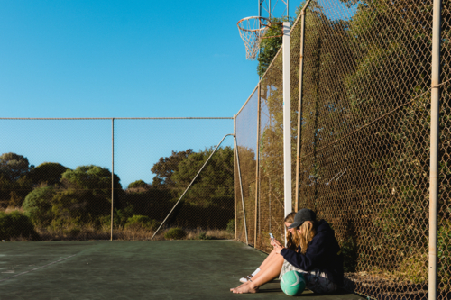 Side view of teenage girls on smart phones with a ball at outdoor netball court - Australian Stock Image