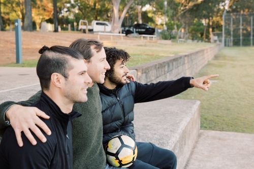Side view photo of 3 smiling interracial man sitting on a field, one is pointing a finger - Australian Stock Image