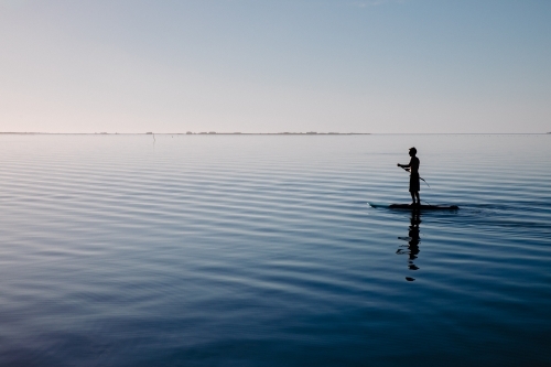Silhouette of a man on a stand-up paddle board on very still water - Australian Stock Image