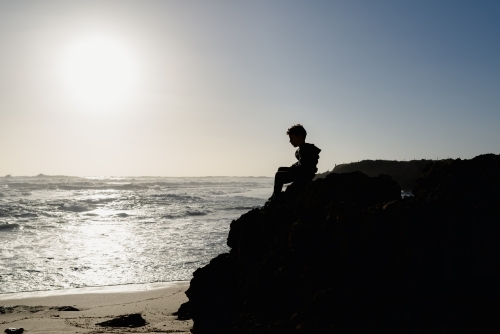Silhouette of a young boy sitting on a rocky ledge on the beach at sunset - Australian Stock Image
