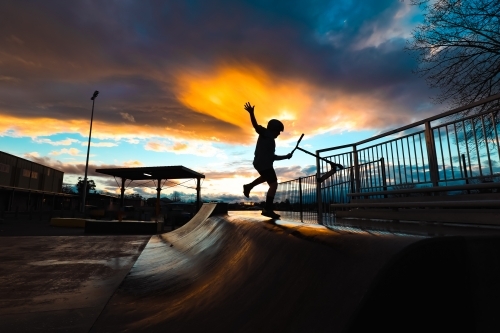 Silhouette of boy riding scooter on skate park ramp at sunset in Mansfield, Victoria - Australian Stock Image