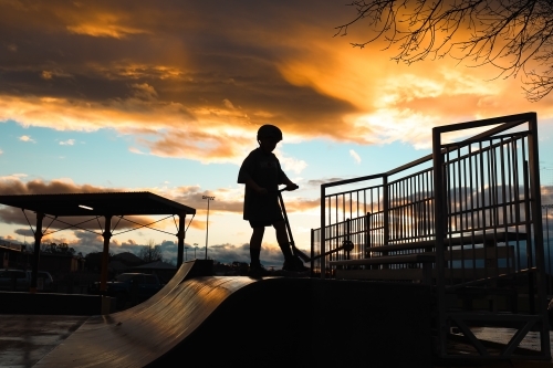 Silhouette of boy riding scooter on skate park ramp at sunset in Mansfield, Victoria - Australian Stock Image