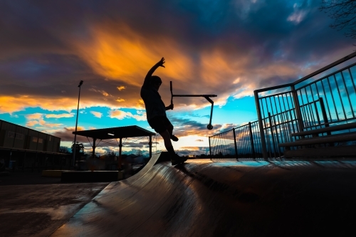 Silhouette of boy riding scooter on skate park ramp at sunset in Mansfield, Victoria - Australian Stock Image