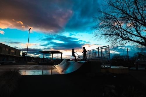 Silhouette of boys riding scooters on skate park ramp at sunset in Mansfield, Victoria - Australian Stock Image