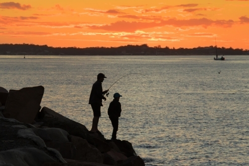 Silhouette of father and son fishing off breakwall at sunset