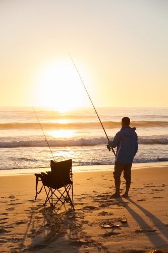 Silhouette of man and camp chair on seashore fishing at sunrise - Australian Stock Image