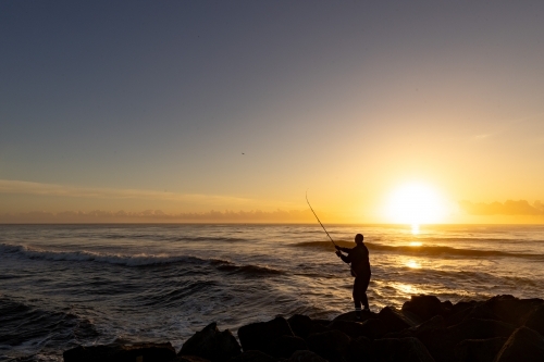 Silhouette of man fishing off rocks at sunrise - Australian Stock Image