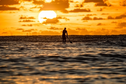 Silhouette of paddler with the large sun rising behind him on the ocean water - Australian Stock Image