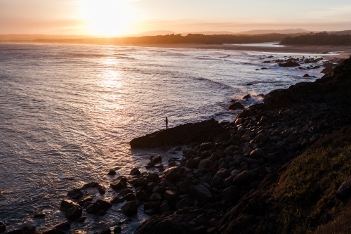 Silhouette of young adult man fishing off the rocks into the ocean at sunset - Australian Stock Image