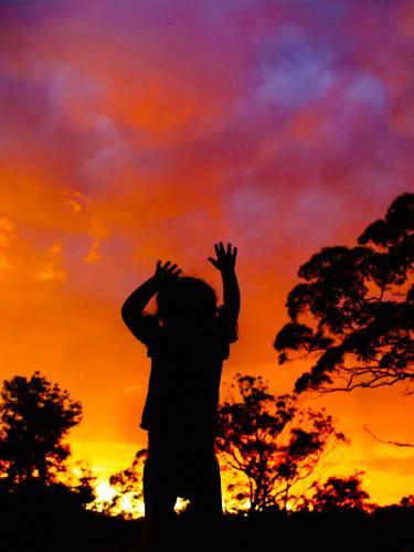 Silhouette of young child reaching up to sky at sunset