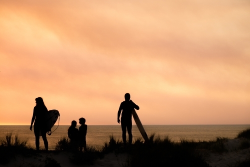 silhouette portrait of surfing family of four overlooking ocean on coastal dune holding surfboards