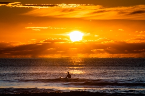 silhouette shot of a man surfing at the beach during sunset/sunrise with orange skies - Australian Stock Image