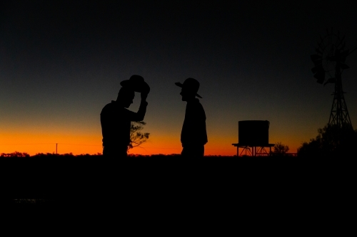 Silhouette shot of two men with hats with one raising his hat during sunset - Australian Stock Image