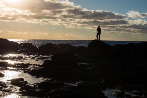 Silhouetted female standing on rocky headland early in the morning - Australian Stock Image