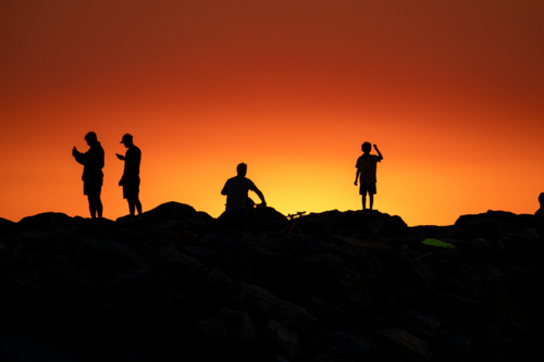 Silhouettes of people on rocks against a vivid orange and yellow sunset sky. - Australian Stock Image