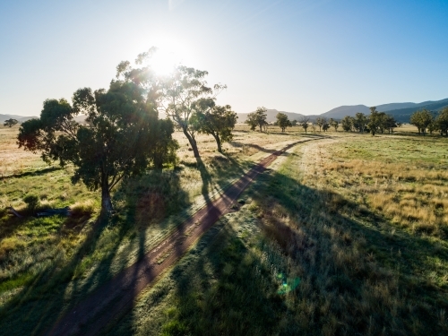 Silver sun shining through trees beside gravel road seen from aerial view on farm - Australian Stock Image