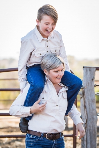 Single mum carrying son on shoulders laughing on farm in drought - Australian Stock Image