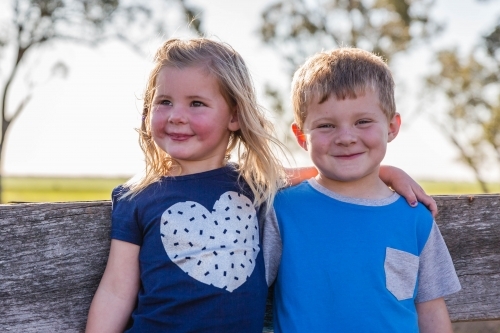 Sister with arm around brother leaning on wooden fence smiling - Australian Stock Image