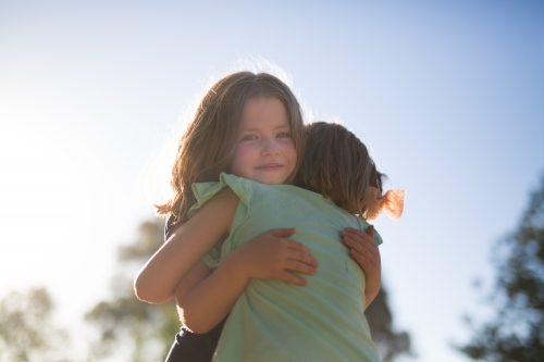 Sisters hugging with sunlight, trees and blue sky in the background - Australian Stock Image
