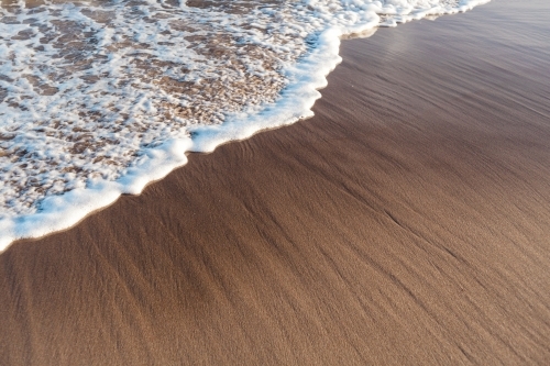 Small, foamy wave washes in against wet textured sand.