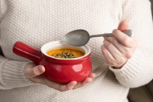 small red bowl of pumpkin soups in lady's hands with spoon - Australian Stock Image