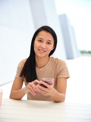 Smiling Asian woman sitting at a table using a phone - Australian Stock Image
