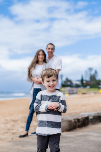 Smiling boy in striped shirt plays at the beach with his parents in the background - Australian Stock Image