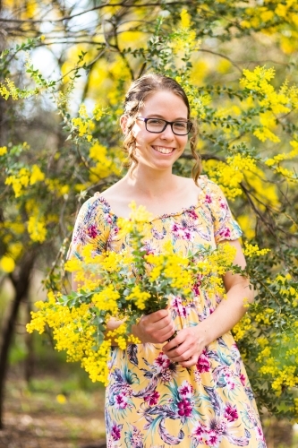 Smiling female in yellow dress with wattle blossom bouquet in native bushland - Australian Stock Image