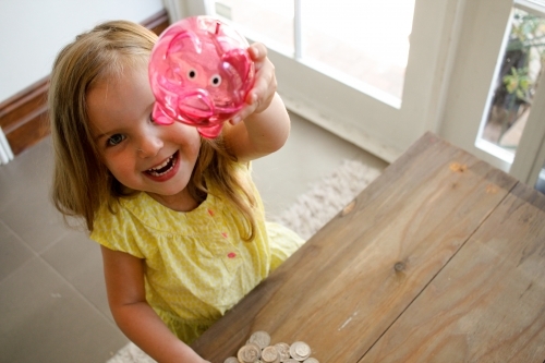 Smiling girl with blonde hair wearing yellow blouse raising a pink transparent piggy bank