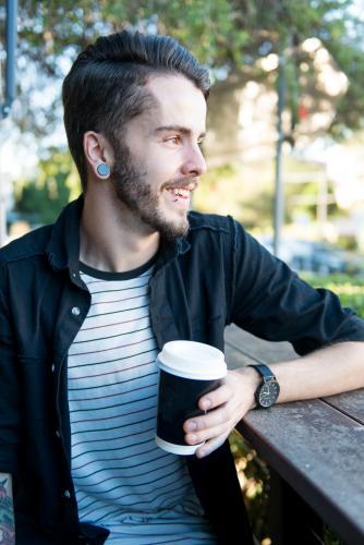 Smiling hipster man sitting outdoors with a takeaway coffee - Australian Stock Image