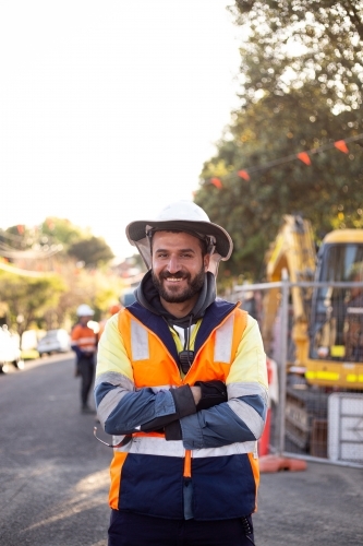 Smiling road worker man with beard wearing orange and yellow high-vis jacket with his arms crossed - Australian Stock Image