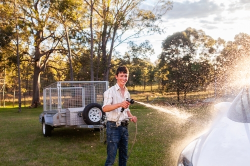 Smiling teen boy washing car spraying vehicle with water - Australian Stock Image