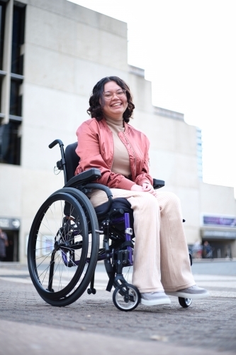 Smiling woman in pink with a disability sitting in a wheelchair outside - Australian Stock Image