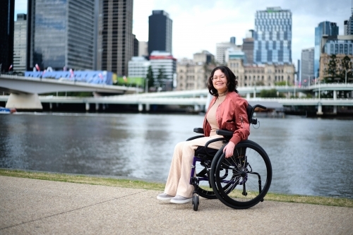 Smiling woman in wheelchair by river with city in background - Australian Stock Image