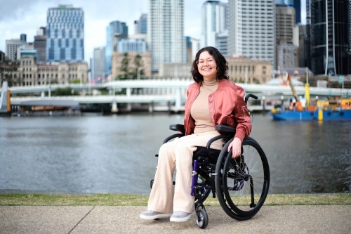 Smiling woman in wheelchair in front of river with city in background - Australian Stock Image