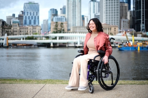 Smiling woman with a disability sitting in a wheel chair in the city with buildings and river - Australian Stock Image