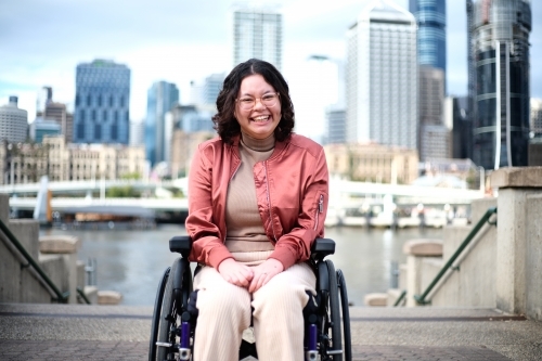 Smiling woman with a disability sitting in a wheel chair with tall buildings behind her - Australian Stock Image