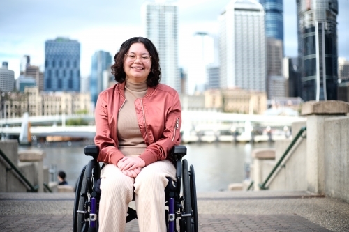 Smiling woman with a disability sitting in a wheel chair with tall buildings behind her - Australian Stock Image