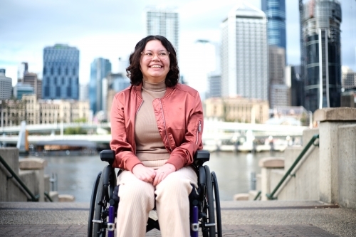 Smiling woman with a disability sitting in a wheel chair with tall buildings behind her