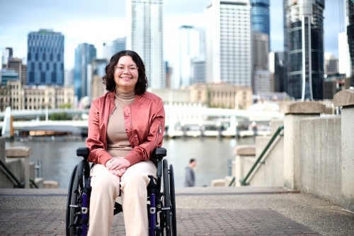 Smiling woman with a disability sitting in a wheel chair with tall buildings behind her - Australian Stock Image