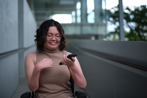 Smiling woman with a disability sitting in a wheelchair indoors holding a mobile phone - Australian Stock Image