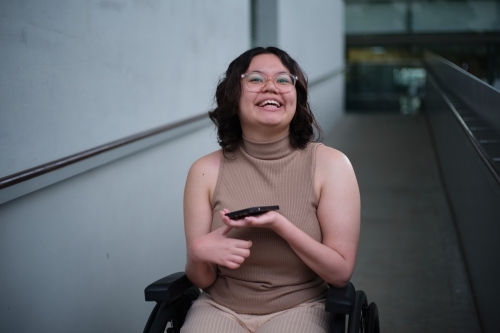 Smiling woman with a disability sitting in a wheelchair indoors holding a mobile phone - Australian Stock Image