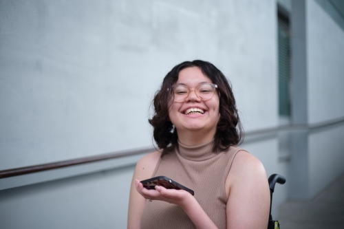 Smiling woman with a disability sitting in a wheelchair indoors holding a mobile phone - Australian Stock Image