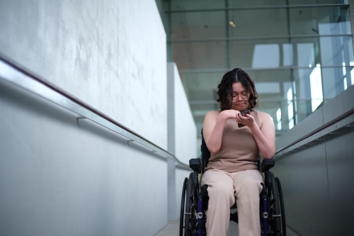 Smiling woman with a disability sitting in a wheelchair looking down at her mobile phone - Australian Stock Image